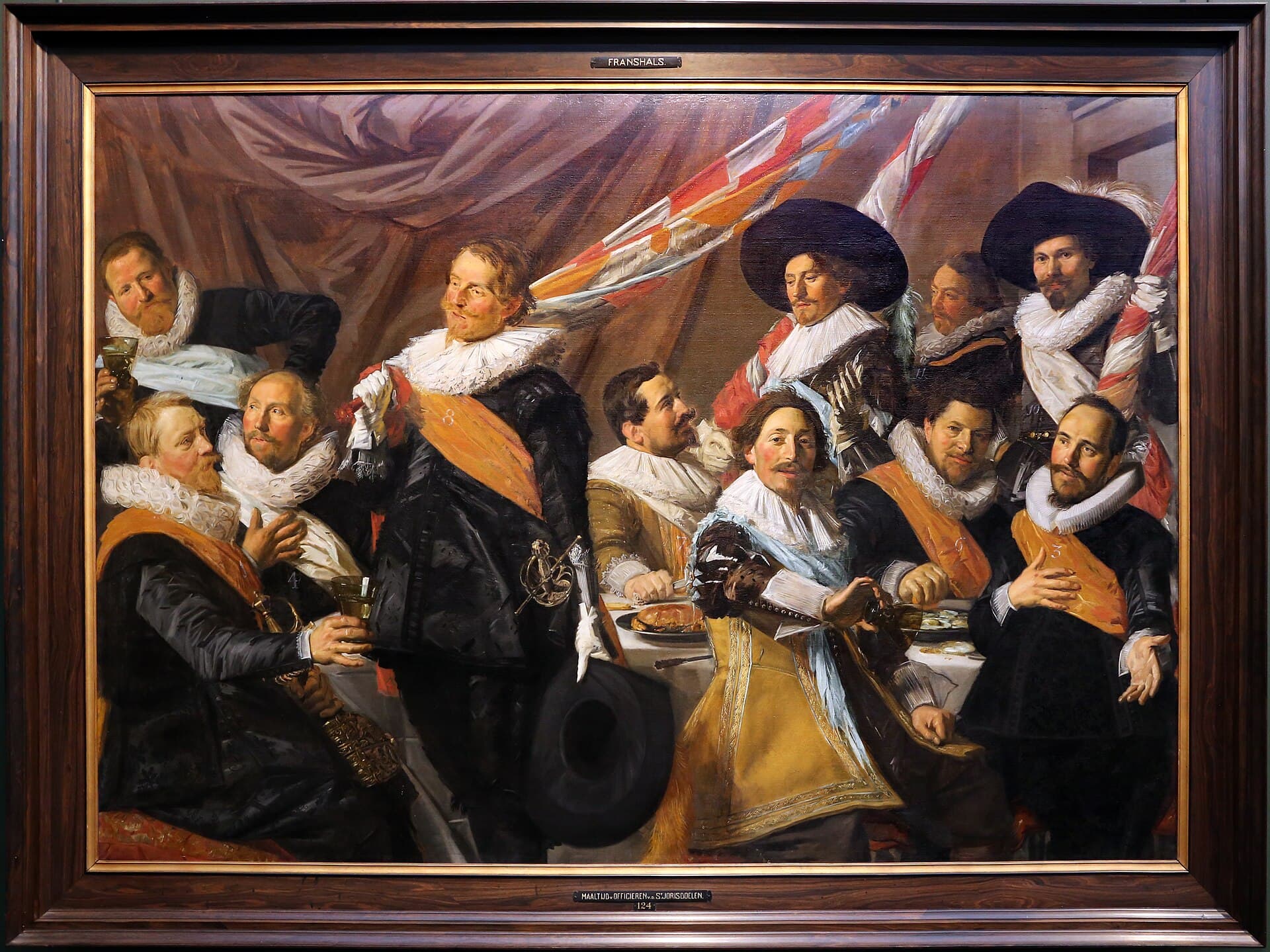 Frans Hals, 1627, olej, Frans Hals Museum w Haarlemie - szeroki portret grupowy kilkunastu mężczyzn stłoczonych wokół długiego stołu nakrytego białym obrusem, na którym stoją talerze i kielichy; wszyscy mają czarne stroje, szerokie białe kryzy pod brodą i barwne szarfy przewieszone przez pierś, jeden unosi kielich, drugi ściska drzewce chorągwi, kilku odwraca się ku sobie w rozmowie, a jeden patrzy poza krawędź obrazu