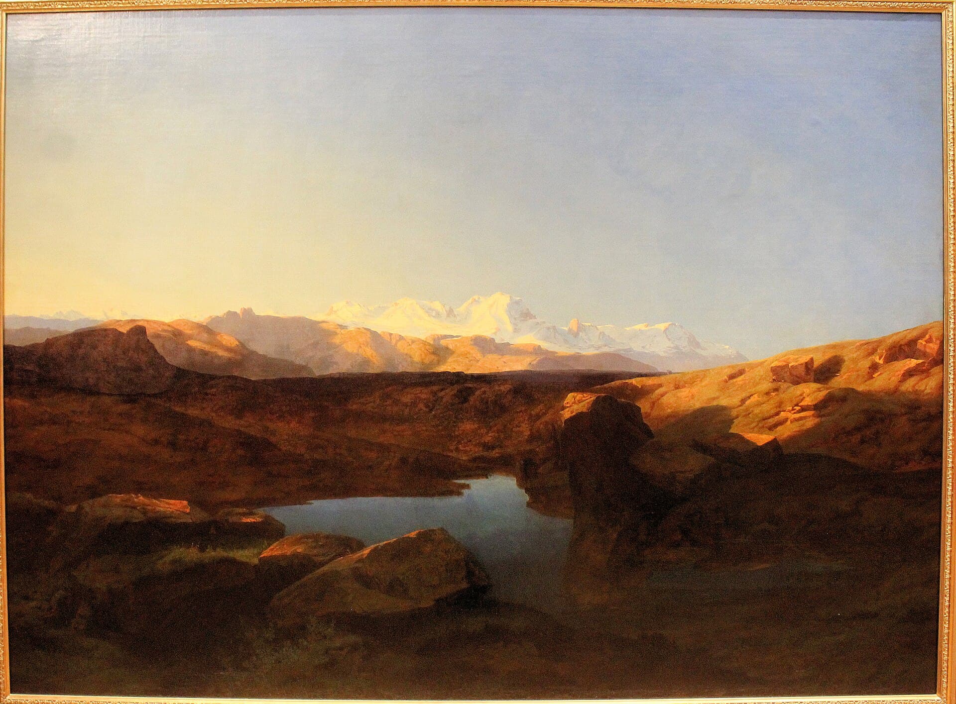 Alexandre Calame, Łańcuch Monte Rosa o wschodzie słońca, XIX wiek, olej - ciemne, brunatne pogórze zajmuje dolne dwie trzecie obrazu, w zagłębieniu między głazami leży małe jezioro, dalej pasmo pogrążone w cieniu, a na horyzoncie ośnieżone szczyty Monte Rosa rozświetlone porannym słońcem pod bladym niebem