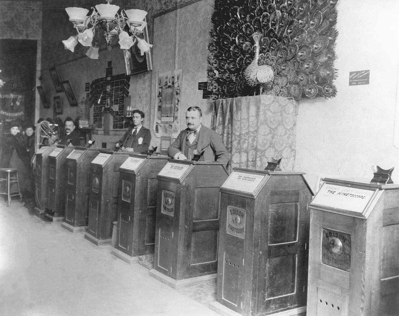 Czarno-biała fotografia wnętrza salonu kinetoskopowego: wzdłuż ściany biegnie ukośnie długi rząd jednakowych drewnianych szaf z napisem Edison Kinetoscope, każda ma na wieku czarny okular i tabliczkę z tytułem; za rzędem stoi kilku mężczyzn w garniturach, przy dalszym końcu dwie kobiety w ciemnych strojach, u sufitu żyrandol z mlecznymi kloszami, a na ścianie rozpostarty paw i oprawione fotografie