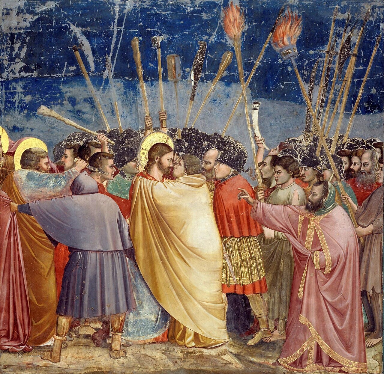 Giotto, Pocałunek Judasza, ok. 1305, kaplica Scrovegnich w Padwie — na tle intensywnie niebieskiego nieba zbity tłum z uniesionymi pochodniami, maczugami i włóczniami; pośrodku mężczyzna w żółtym płaszczu zagarnia nim postać z aureolą i przybliża twarz do jej twarzy, oboje patrzą sobie w oczy, z lewej ktoś zarzuca komuś płaszcz na głowę, z prawej stoi tyłem mężczyzna w różowej szacie z wyciągniętą przed siebie ręką
